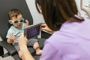 A young boy wearing sunglasses is looking at a tablet while sitting on a chair, and a woman in a purple shirt is holding the tablet.