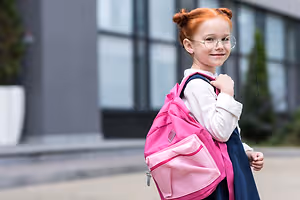 A young girl with red hair is wearing a white long-sleeve shirt and a blue skirt, carrying a pink backpack, and smiling in front of a blurry background with a glass wall and plants.
