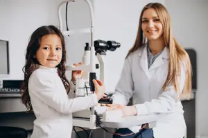 A young girl and a woman in a white lab coat are standing in front of an eye examination machine.