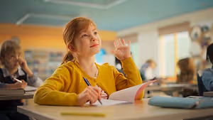 A girl sitting at a desk in a classroom is holding a pen and a book and raising her hand