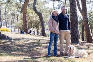 A couple and their dog are standing in a wooded area.