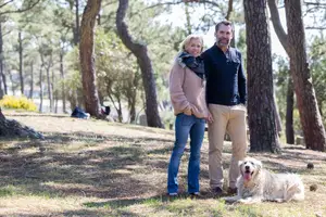 A couple and their dog are standing in a wooded area.