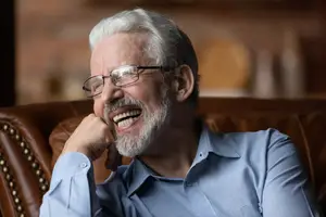 An older man wearing glasses and a blue shirt is sitting on a brown leather couch, smiling and looking to the right with his right hand on his chin.