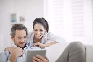 A man and woman sitting on a couch with the woman looking at a tablet while the man holds it.