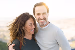 A smiling man and woman are standing close to each other with their arms around each other.