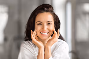 A woman is smiling and posing for a picture while holding her cheeks with both hands.