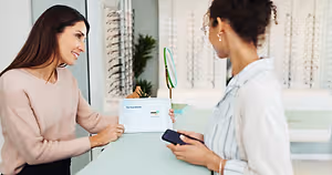 A woman in a white shirt is holding a phone and looking at a tablet while a woman in a pink shirt smiles and holds a tablet in an office. Behind them is a plant and a mirror.