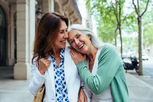 Two women standing and laughing together on a sidewalk, one holding a handbag and the other hugging the other woman