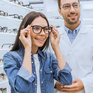 A woman trying on glasses in a store with an optician in the background