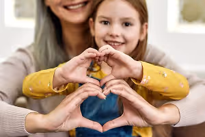 A woman and a young girl are smiling and making a heart shape with their hands