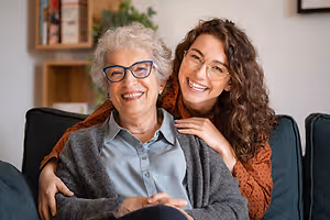 An elderly woman and a young woman are sitting on a couch, smiling and posing for a photo.