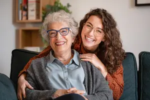 An elderly woman and a young woman are sitting on a couch, smiling and posing for a photo.
