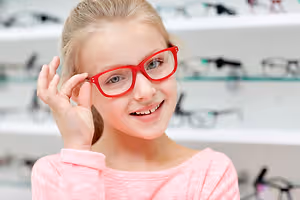 A young girl wearing red glasses is smiling and posing for a photo in an optics shop.