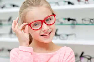 A young girl wearing red glasses is smiling and posing for a photo in an optics shop.