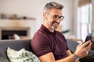 A smiling man with glasses and a watch is sitting on a couch, holding a cell phone in his right hand, and probably talking to someone.