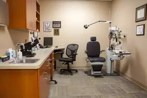 An indoor optometrist's office with a desk, monitors, chairs, and a microscope on a counter.