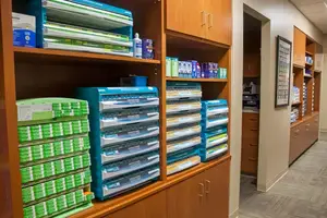 An office or clinic supply room with organized shelves of neatly stacked binders and boxes, and a framed picture on the wall.