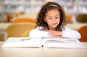 A girl with curly hair sits at a table in a library reading a book