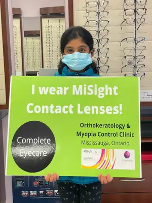 A girl is holding a green sign that says I wear MiSight Contact Lenses in an optical shop.