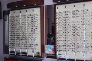 A display of eyeglasses in a store with a mirror reflecting a person wearing a mask and a child wearing a hat