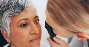 A woman with gray hair is having her eyes examined by another woman who is holding a device