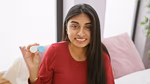 A smiling woman in a red shirt is sitting on a bed and holding a blue and white contact lens.