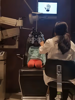 A girl in a green sweater is sitting on a chair while a woman in a white lab coat is doing an eye exam on her in a clinic