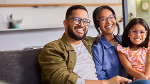 A family of three sitting on a couch, smiling, with a girl in a pink floral dress and glasses, a man in a green jacket, and a woman in a blue shirt, and a cabinet with a potted plant behind them.