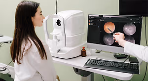 A woman looking into an eye examination machine while a doctor points to an image on the screen