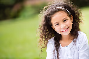 A young girl with curly hair is smiling and looking at the camera, wearing a white shirt and a headband, sitting on a green grass field with a blurry background.