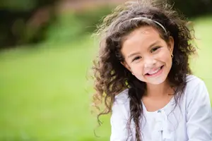 A young girl with curly hair is smiling and looking at the camera, wearing a white shirt and a headband, sitting on a green grass field with a blurry background.