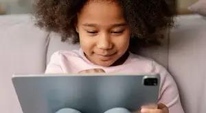 A girl is sitting on a couch while holding a tablet with both hands and smiling. She has curly hair and is wearing a pink shirt and blue jeans. She is probably playing a game on the tablet. Behind her is a white couch with a pillow on it.
