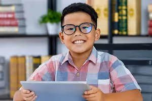 A smiling boy with glasses is holding a tablet in a room with bookshelves and a plant in the background.