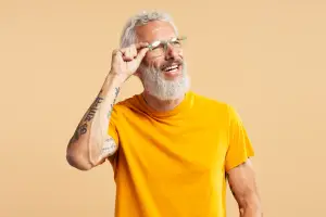 A man wearing a yellow t-shirt and glasses is smiling and looking at the camera with his hand on his face