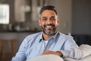 A man with a beard is smiling and sitting on a couch with his hand on the armrest in a room.