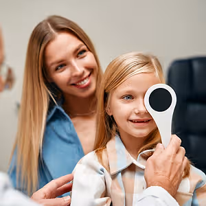 A young girl is having her eyes examined by a doctor in a clinic, with her mother standing beside her, all smiling.
