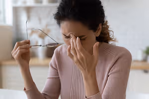 A woman sits at a table holding a pair of glasses, touching her forehead, and looking tired
