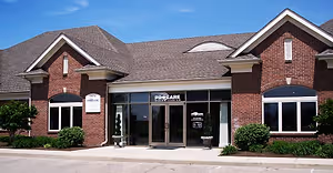 A modern brick building with a gray roof, glass doors, and windows, named ProCare, is set against a blue sky with clouds. It has a paved entrance and greenery around it.