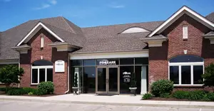 A modern brick building with a gray roof, glass doors, and windows, named ProCare, is set against a blue sky with clouds. It has a paved entrance and greenery around it.