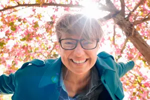 A smiling woman in a teal coat stands under a cherry blossom tree, with sunlight shining through the branches