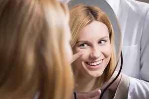 A woman in a white coat is holding a mirror and looking at her face with a smile on her face.