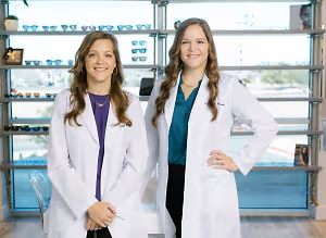 Two women in white lab coats standing in front of a shelf with glasses