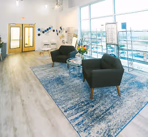 Interior of a modern waiting room with glass windows, wooden flooring, and a blue rug with two chairs and a table with flowers. There is also a potted plant on the left side.