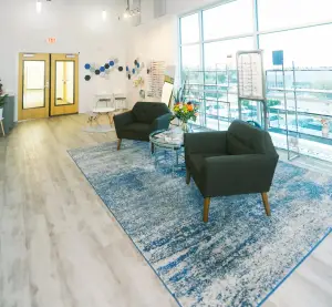 Interior of a modern waiting room with glass windows, wooden flooring, and a blue rug with two chairs and a table with flowers. There is also a potted plant on the left side.