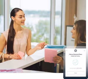 Two women, one smiling and holding a pink card, are sitting at a desk with a glass window and a lake in the background. The woman on the left is wearing a pink blouse, a necklace, and a bracelet, while the woman on the right is wearing a black top and has a bag. There is a smartphone displaying a transaction with a checkmark, and a watermark with the text Cherry Payment Plans.