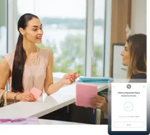 Two women, one smiling and holding a pink card, are sitting at a desk with a glass window and a lake in the background. The woman on the left is wearing a pink blouse, a necklace, and a bracelet, while the woman on the right is wearing a black top and has a bag. There is a smartphone displaying a transaction with a checkmark, and a watermark with the text Cherry Payment Plans.