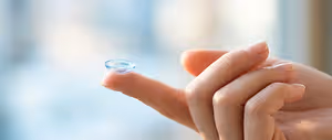 A person's hand holding a blue contact lens, likely for vision correction, against a blurred background.