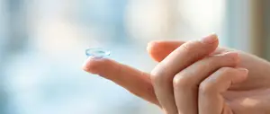 A person's hand holding a blue contact lens, likely for vision correction, against a blurred background.