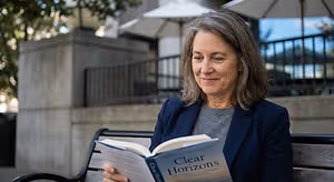 An older woman in a gray shirt and blue blazer sits on a bench outdoors and reads the book Clear Horizons