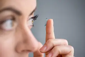 A woman in a gray background puts a contact lens on her eye.